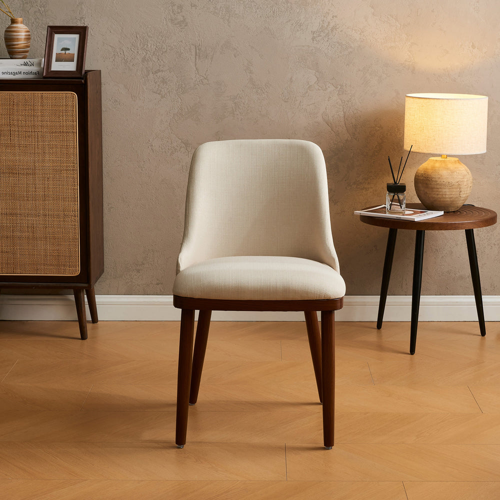 Front view of Modern Upholstered Dining Chair in cream linen with tapered walnut legs by a lamp