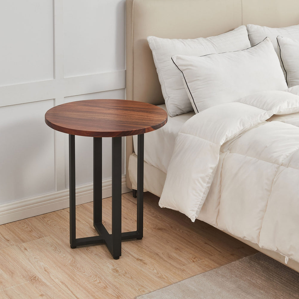 Round End Table in walnut with black metal X-base beside an upholstered bed