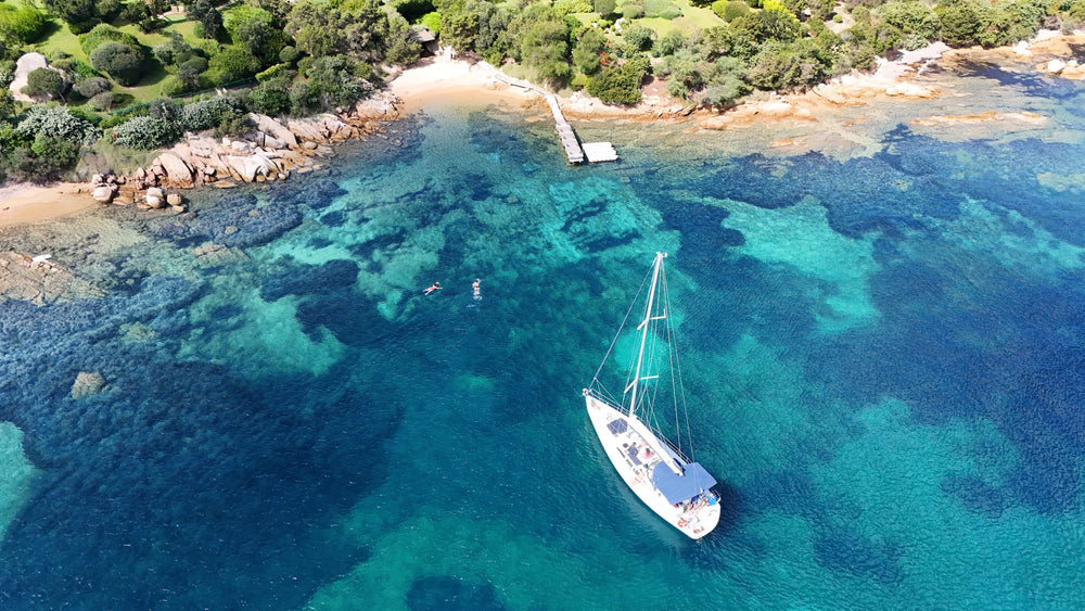 Aerial drone photo of a white sailboat anchored in clear turquoise water beside a rocky green cove